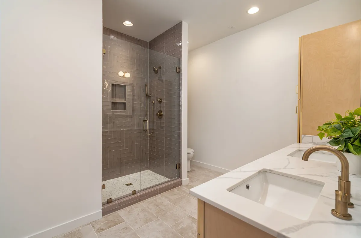 A modern bathroom with a luxurious glass shower enclosure, marble countertop, wood cabinetry, and a potted plant for a touch of freshness. The serene palette and stylish fixtures emphasize a spa-like, rejuvenating experience.