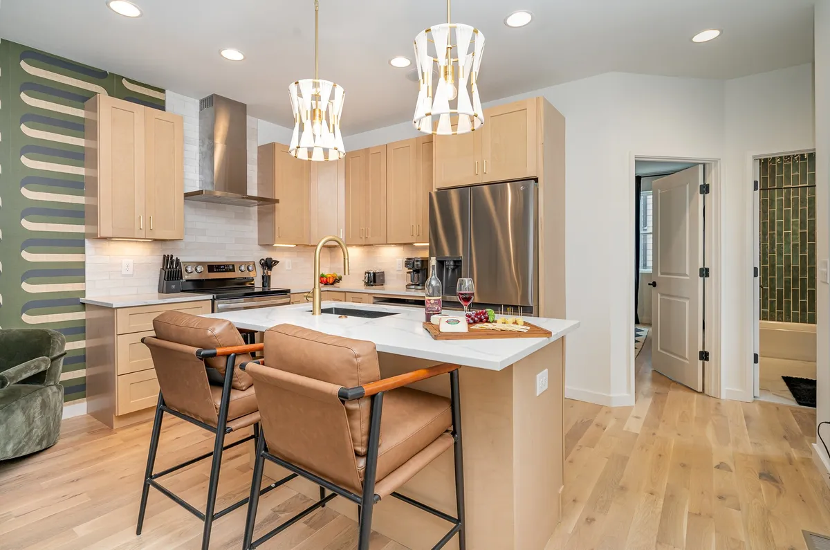 A warm, welcoming kitchen scene with a kitchen island, bar stools, wall art, appliances, and elegant lighting. The space is designed for interaction—hosting dinner parties, baking, or sharing a glass of wine after a long day.