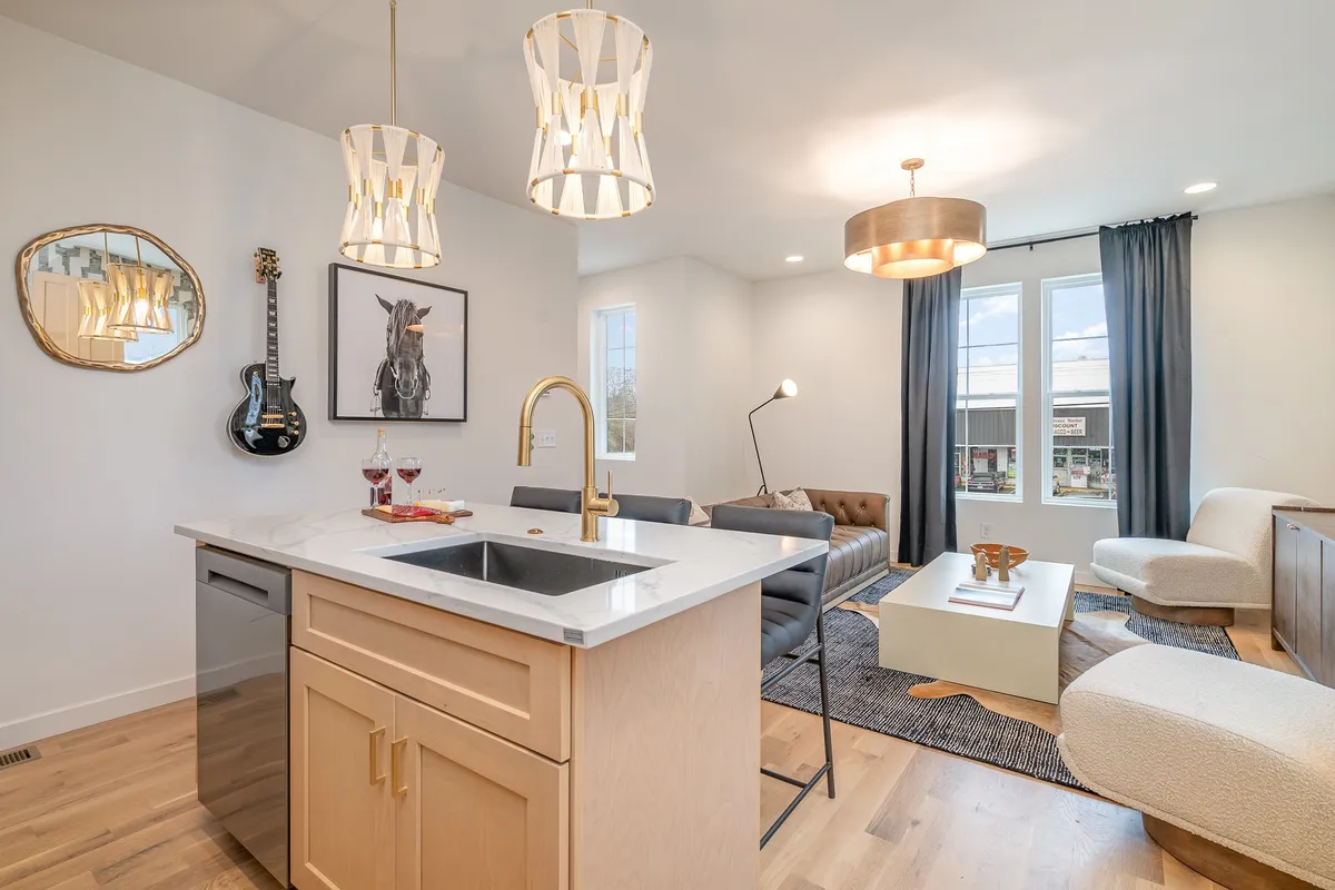 A bright, contemporary kitchen and living area with a central island, chandelier lights, wall-mounted guitar, horse artwork, and a modern sofa. The space feels both functional and stylish, perfect for preparing meals or gathering with friends and family.