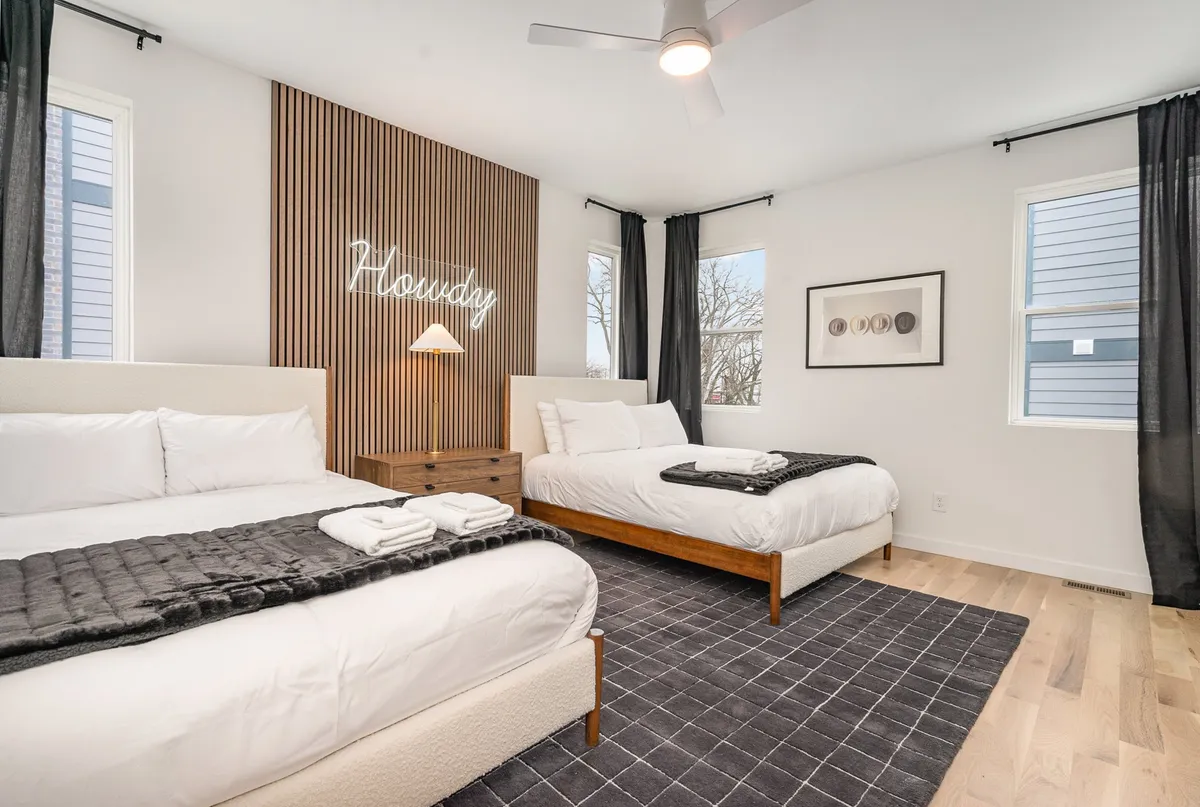 A cozy bedroom featuring two beds, a neon sign, wooden accent wall, and black curtains. The design is modern, with an emphasis on comfort and inviting rest.