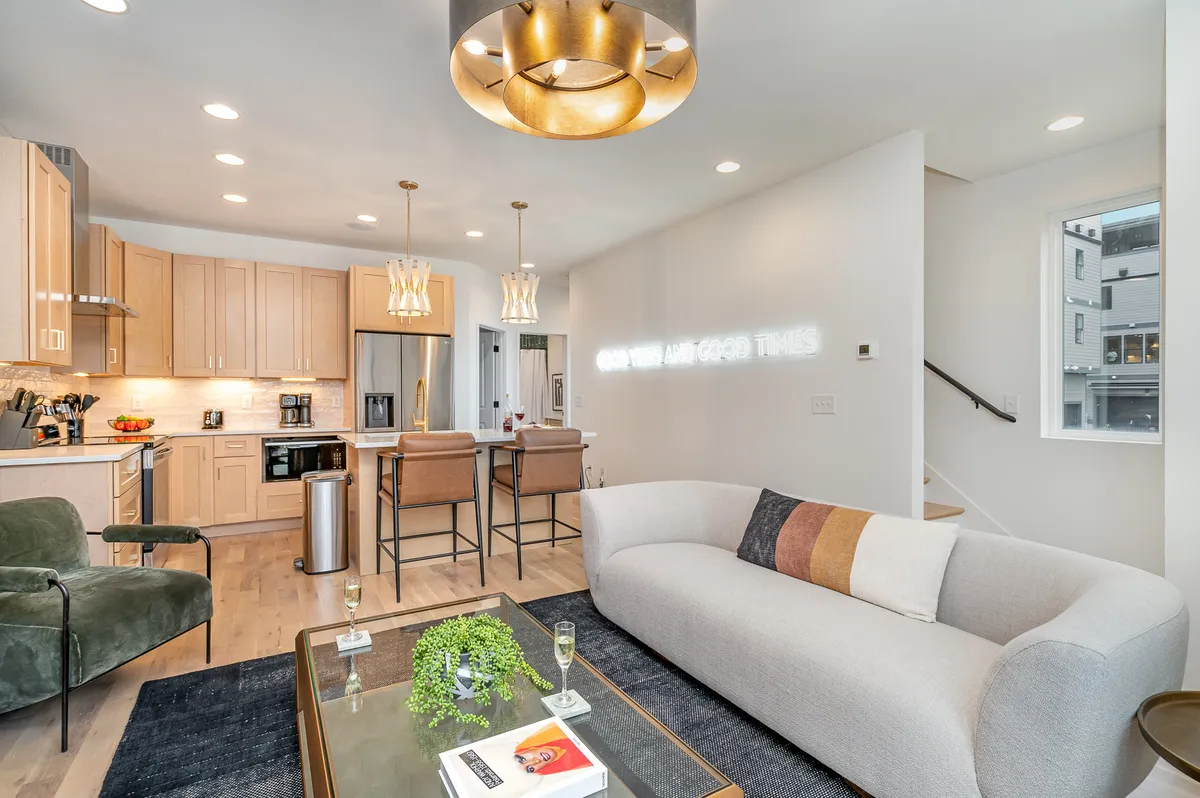 This image presents an open-concept living area featuring a sophisticated sofa, a modern kitchen island, decorative lighting fixtures, and artful wall decor. The space seamlessly blends lounging and kitchen areas, perfect for gatherings, conversations, and shared meals.