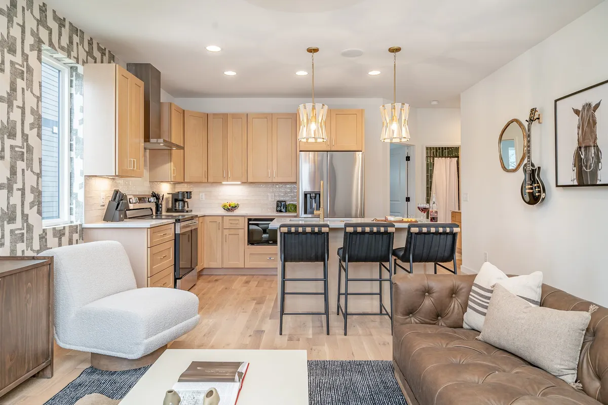 This image showcases a welcoming interior featuring a kitchen island with bar stools, a comfortable sofa with decorative pillows, stylish wall art, and modern light fixtures. The open-plan layout and inviting decor evoke a sense of homey comfort and practicality.