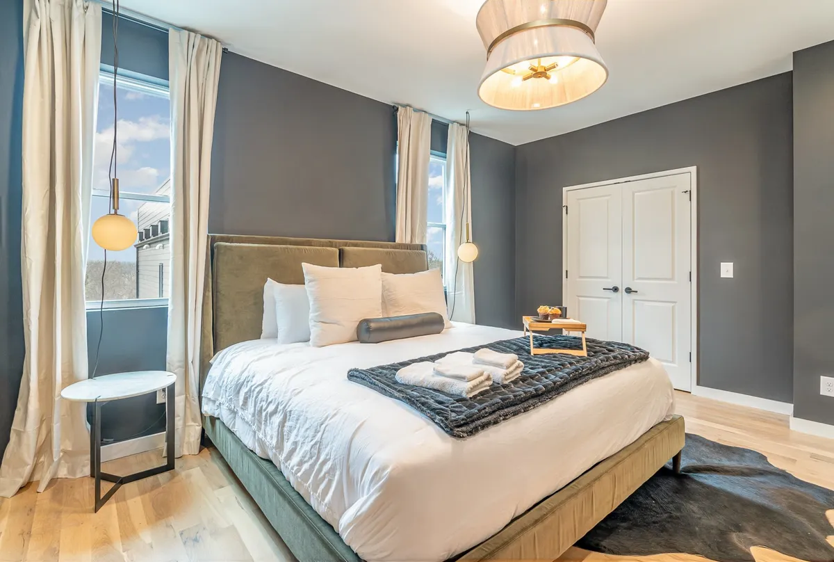 A tranquil bedroom with a large bed dressed in plush bedding, framed by elegant curtains and a stylish side table with decorative accents. The modern light fixture and soft color palette create a restful, inviting atmosphere ideal for relaxation and rejuvenation.