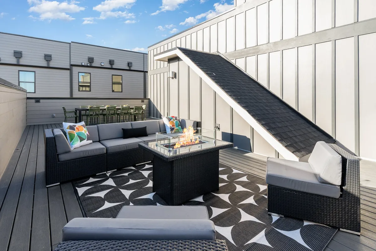 An inviting outdoor rooftop seating area centered around a glowing fire pit, with a decorative rug and plush seats creating a cozy yet open atmosphere. Ideal for casual conversations and fireside gatherings as the evening sets in.