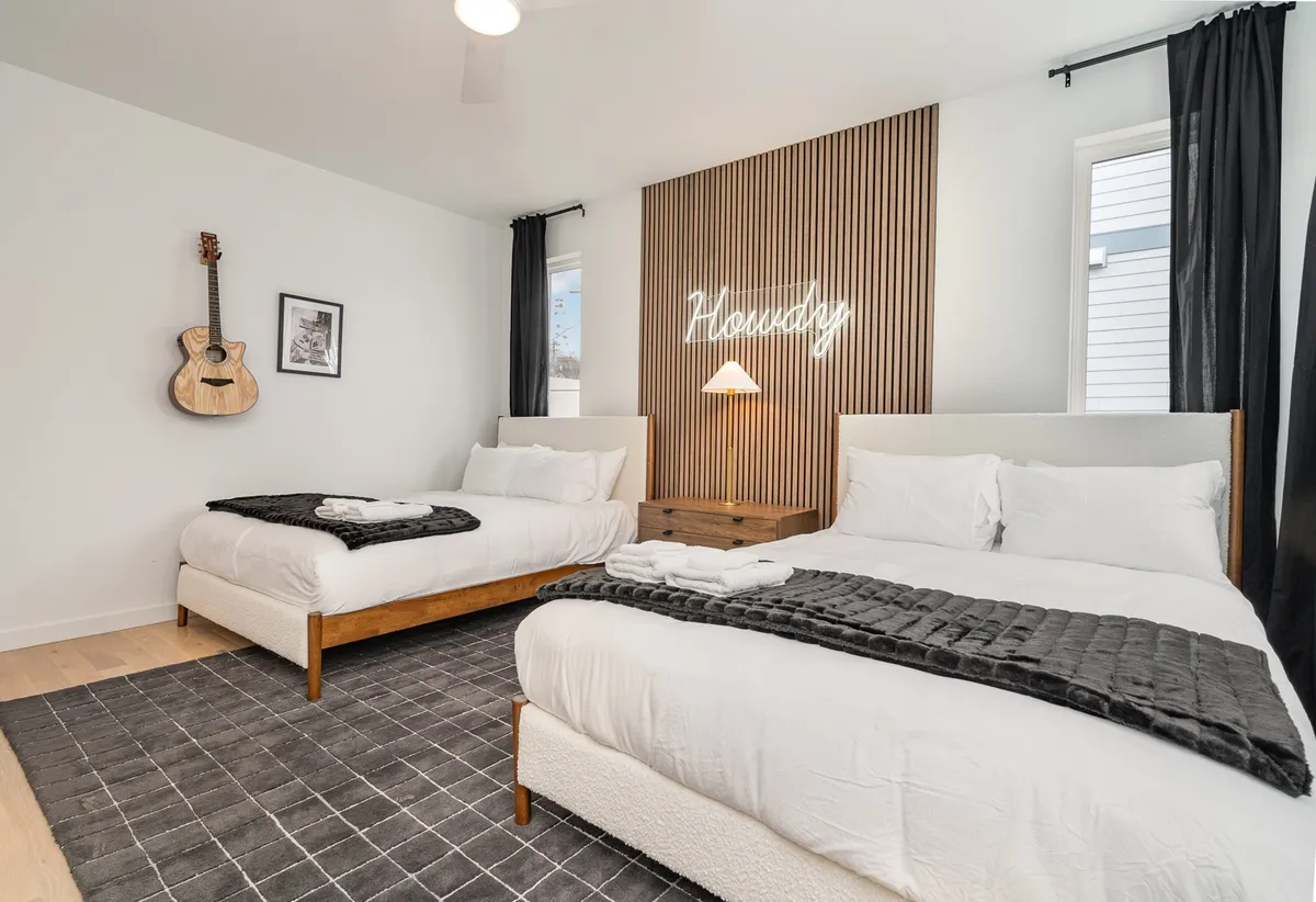 A cozy bedroom featuring two beds with plush bedding, a decorative neon sign, and an acoustic guitar mounted on the wall. The setting is both stylish and welcoming, with touches of personality and a layout that works well for friends or family sharing a space.